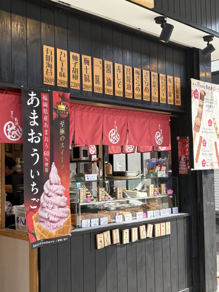 太宰府参道にある寺子屋本舗の店頭。串ぬれおかきや、福岡県産あまおうソフトクリームの看板が並び、食べ歩きグルメが楽しめる様子