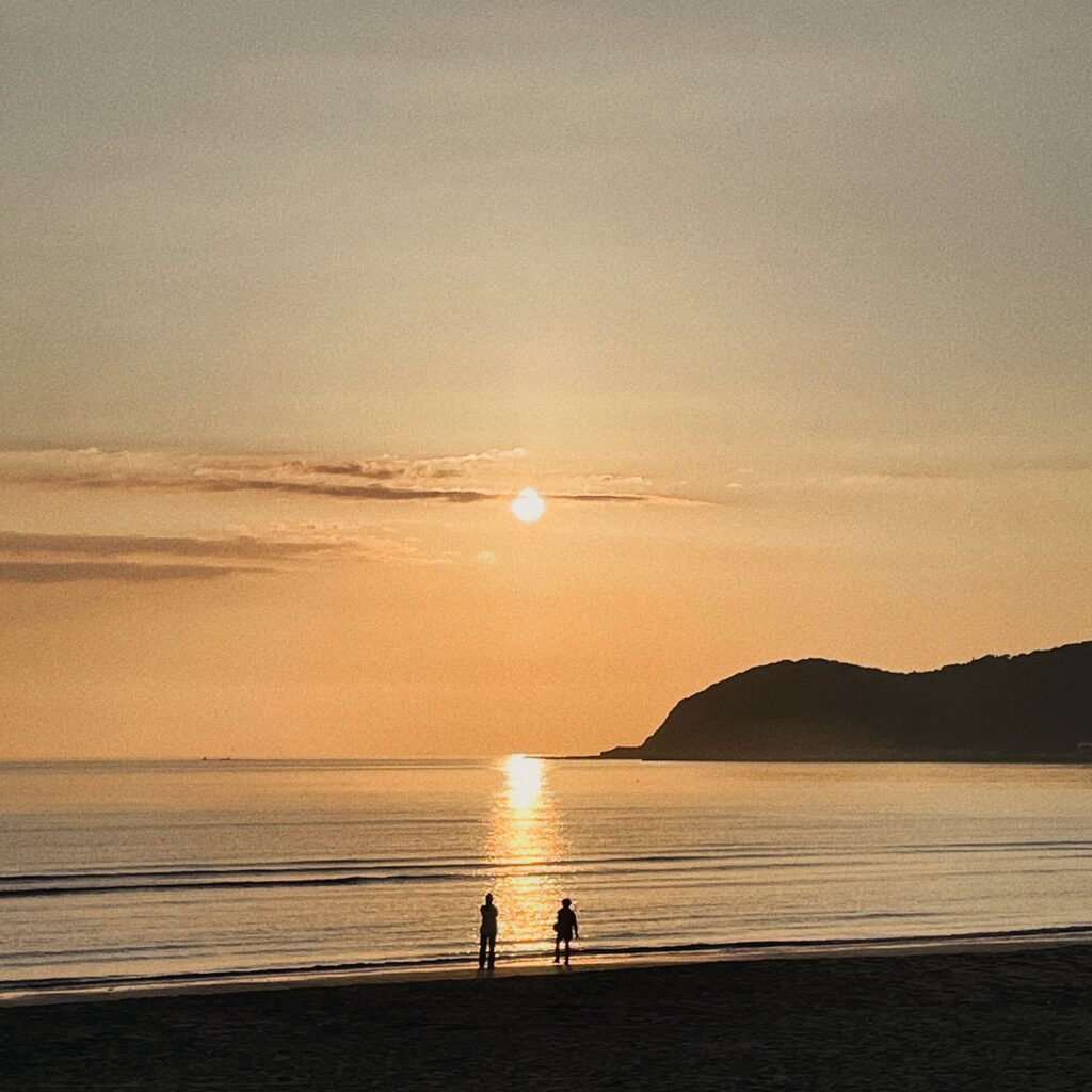 夕日に照らされた福間海岸。オレンジ色に染まる海と、散歩する人のシルエット