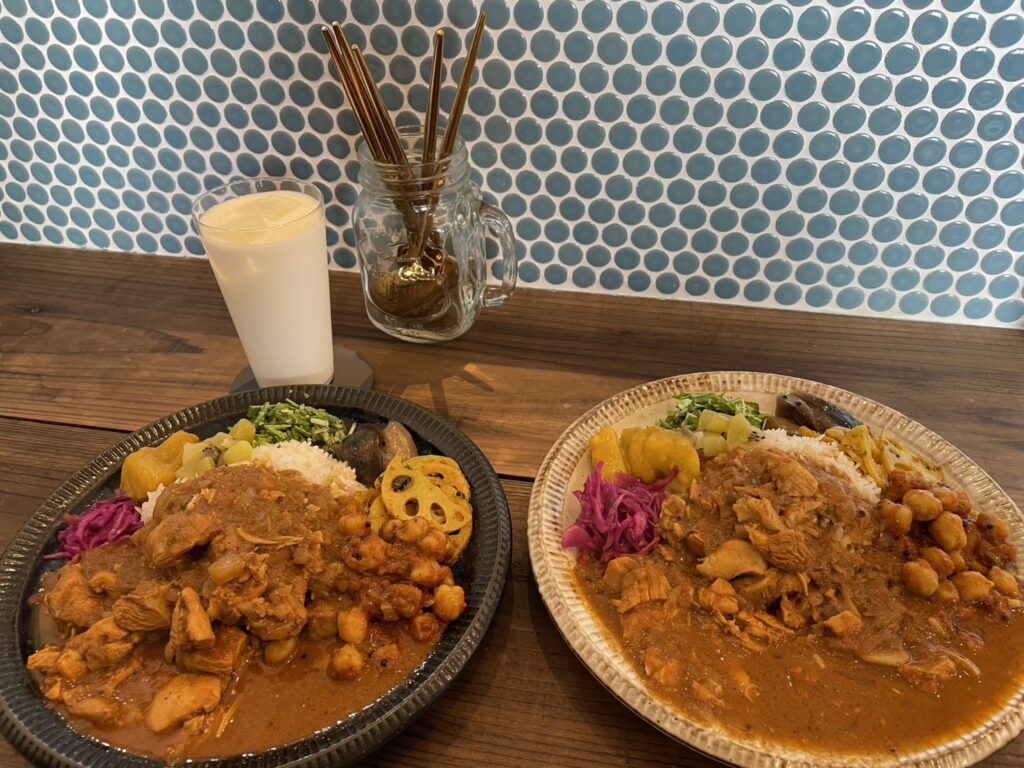 マイコのカレーの本日のプレート２皿の画像。チキンのトマトカレーをメインに、ひよこ豆のトマト煮やナスの甘酢炒め、紫キャベツのマリネなどが乗っている。
