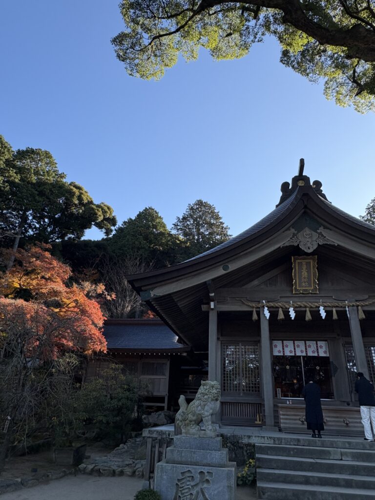 竈門神社の本殿と、境内の赤く色づいた紅葉。青空の下で参拝する人の様子。